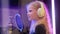 Young blonde girl singer sings into microphone in white headphones in professional recording studio in multicolored rays
