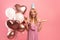 Young blonde lady with festive balloons, birthday cap and party blower celebrating special occasion on pink background