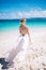 Young blonde long hair bride in white dress with open back standing on the beach. Tropical turquois ocean on the background.