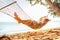 Young blonde longhaired woman relaxing in hammock hinged between palm trees on the sand beach