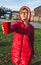 Young blonde serious girl in red clothes standing on a street gives a cup with water