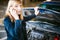 Young blonde stands near car with raised engine compartment hood