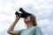 Young blonde woman bird watcher in cap and blue t-shirt looking through binoculars at cloudy sky in summer forest ornithological