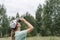 Young blonde woman bird watcher in cap and blue t-shirt looking through binoculars at cloudy sky in summer forest ornithological