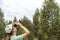 Young blonde woman bird watcher in cap and blue t-shirt looking through binoculars at cloudy sky in summer forest ornithological