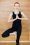 Young blonde Woman in a black suit working out indoors, doing yoga exercise on mat, standing in Vrksasana Posture, Tree Pose, full