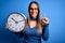 Young blonde woman with blue eyes holding big minute clock over isolated background surprised with an idea or question pointing