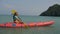 Young blonde woman in blue swimsuit rows pink plastic canoe along azure sea bay past island with palms under blue sky at