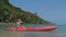 Young blonde woman in blue swimsuit rows pink plastic canoe along azure sea bay past island with palms under blue sky at