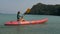 Young blonde woman in blue swimsuit rows pink plastic canoe along azure sea bay past island with palms under blue sky at