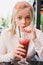 Young blonde woman with freckles wondering and drinking red coctail with berries and mint in outdoor restaurant