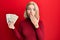 Young blonde woman holding 100 danish krone banknotes covering mouth with hand, shocked and afraid for mistake