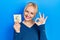 Young blonde woman holding euro symbol reminder doing ok sign with fingers, smiling friendly gesturing excellent symbol