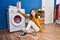 Young blonde woman sitting on floor turning washing machine on at laundry room