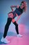 Young blonde woman in velour blue sport wear and black stockings seductively posing on camera in studio. Pink and blue