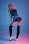 Young blonde woman in velour blue sport wear and black stockings seductively posing on camera in studio. Pink and blue