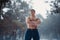 Young bodybuilder with bare torso stands with arms crossed in winter misty forest.