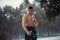 Young bodybuilder with bare torso wipes his body by snow in forest in winter.