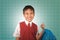 Young boy 7 or 8 years old in vest and necktie uniform holding bag smiling cheerful in front of green chalkboard excited about