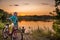 A young boy with a bicycle stands by the lake and admirs the evening sunset.  A friend stands in the distance with a butterfly net
