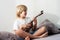 Young boy tuning ukulele at home. Blond haired boy sitting on couch playing acoustic guitar