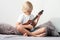 Young boy tuning ukulele at home. Blond haired boy sitting on couch playing acoustic guitar