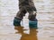 Young boy is washing rubber boots in muddy water of pond. Dirty sand