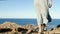 Young, brave woman standing on top of rock on a summer day watching epic sea landscape with dress moving in the wind , back view