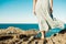 Young, brave woman standing on top of rock on a summer day watching epic sea landscape with dress moving in the wind , evocative