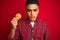 Young brazilian man holding a cookie standing over isolated red background with a confident expression on smart face thinking
