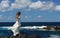 Young bride in light white wedding dress standing on windy sea rock shore. Flowers in hand. Ocean waves, splash of water