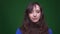 Young brunette female student being embarrassed and shy watches into camera on green background.