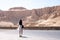 Young brunette female in white dress walks in front of part of the Mortuary temple of the Queen Hatshepsut Dayr el