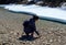 Young brunette girl with backpack raising a stone squatting by a river melting from ice in spring. Solo traveller.