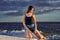 A young brunette girl in a black T-shirt and denim shorts kneels on the sandy shore near the water`s edge.