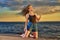 Young brunette girl in a black T-shirt and denim shorts kneels on the sandy shore by the sea.