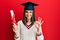 Young brunette girl holding graduate degree diploma doing ok sign with fingers, smiling friendly gesturing excellent symbol