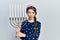 Young brunette girl holding menorah hanukkah jewish candle in shock face, looking skeptical and sarcastic, surprised with open