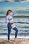 A young brunette girl in a white blouse and blue jeans walks barefoot along the sandy seashore near the water`s edge.