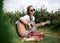 Young brunette hippie woman, wearing boho style clothes, sitting on green grass, holding guitar. Indie musician relaxing on green