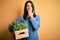 Young brunette woman with blue eyes holding wood box garden of fresh natural plants cover mouth with hand shocked with shame for