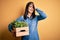 Young brunette woman with blue eyes holding wood box garden of fresh natural plants stressed with hand on head, shocked with shame