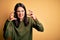 Young brunette woman with blue eyes wearing green casual sweater over yellow background Shouting frustrated with rage, hands