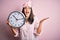 Young brunette woman with blue eyes wearing pajama holding big clock at night time very happy and excited, winner expression