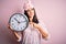 Young brunette woman with blue eyes wearing pajama holding big clock at night time very happy pointing with hand and finger