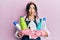 Young brunette woman with curly hair wearing cleaner apron holding cleaning products making fish face with mouth and squinting