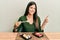 Young brunette woman eating sushi sitting on the table smiling happy pointing with hand and finger to the side