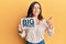 Young brunette woman holding big prize paper smiling happy and positive, thumb up doing excellent and approval sign