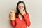 Young brunette woman holding bottle with boiled potatoes covering mouth with hand, shocked and afraid for mistake