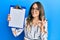 Young brunette woman holding clipboard with blank space doing ok sign with fingers, smiling friendly gesturing excellent symbol